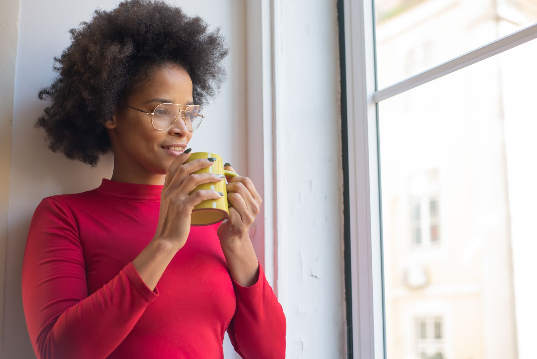 African American woman in a red sweater holding a cup and gazing out of a window with a peaceful expression, illustrating Understanding the Importance of Breaks