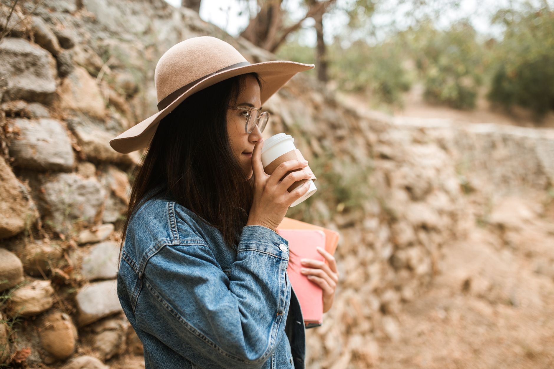 Woman wearing denim jacket and sun hat holding coffee cup in a park, illustrating Overcoming Barriers to Taking Breaks