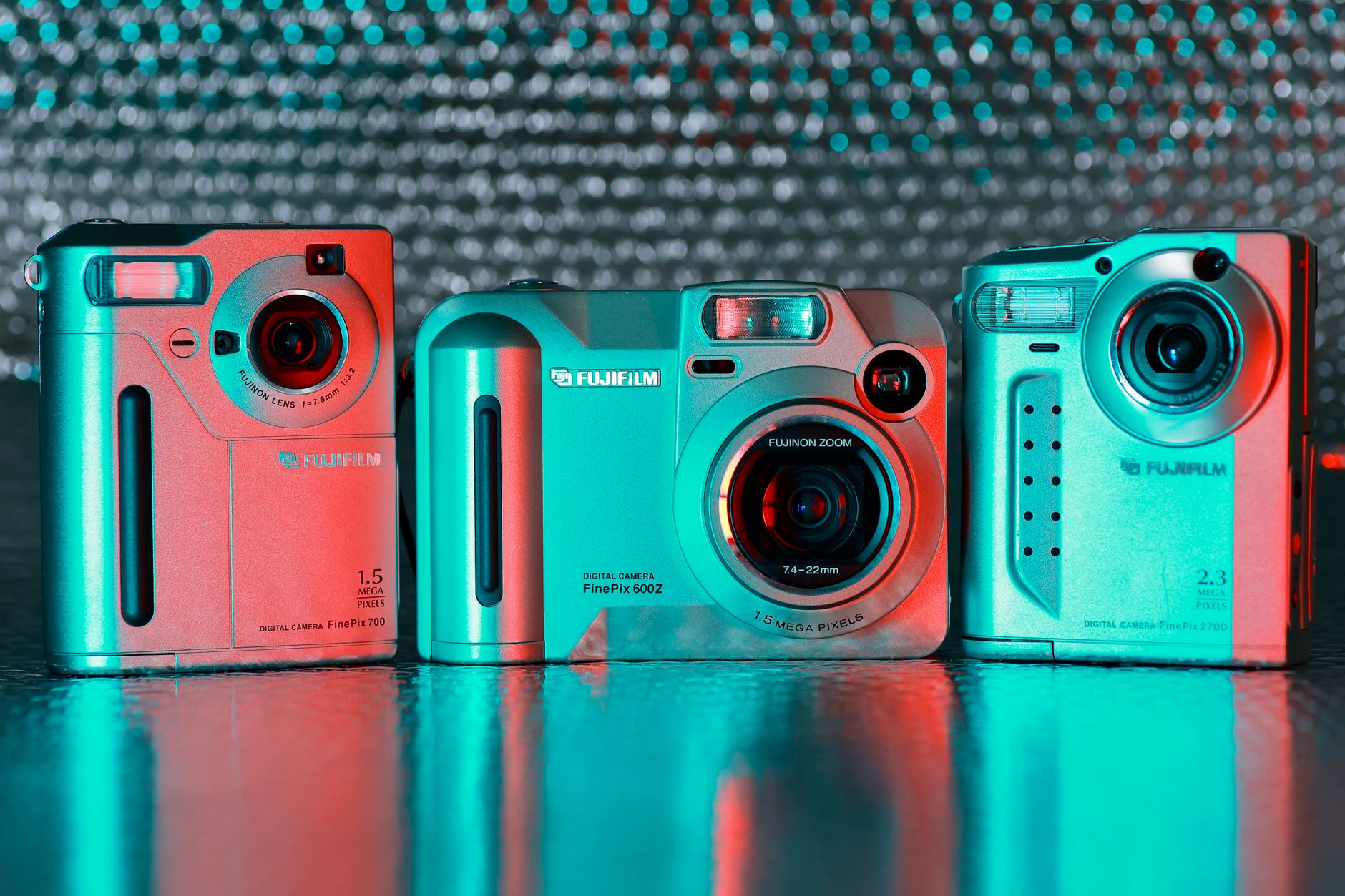 Three vintage Fujifilm cameras with vibrant red and teal neon lighting on a reflective surface, illustrating Check three things before you buy any old digital camera