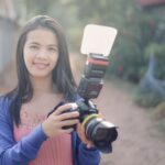 Portrait of a woman smiling and holding a digital camera with flash outdoors, illustrating The Digicam Trend Is Real, but You Don't Need a $1,600 Camera to Get the Look