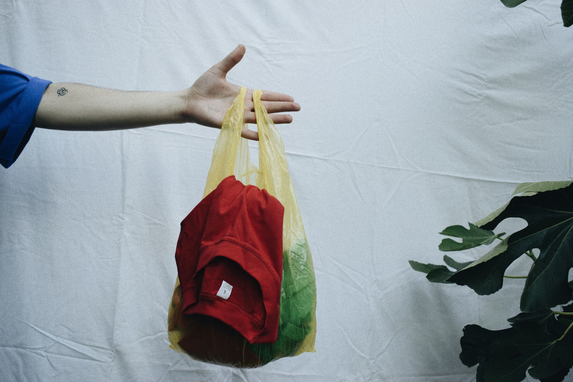 A hand holds a yellow plastic bag containing clothes on a simple white background, illustrating Clothes that sit unsold aren't helping the planet either