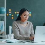 A woman using a laptop in a modern office setting with notebooks and sticky notes on the wall, illustrating Freelancing 101: How to Launch Your Creative Career as a Solopreneur