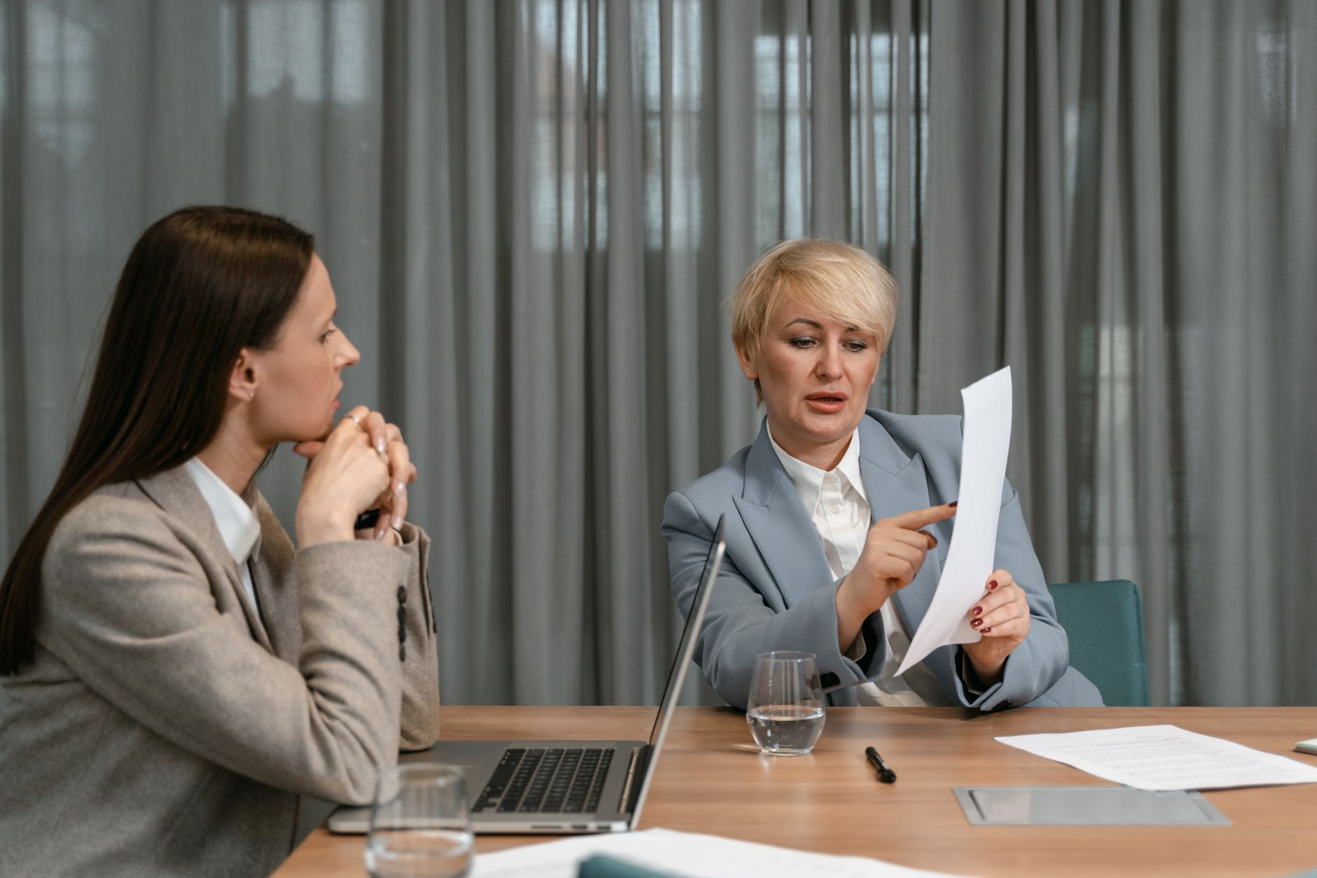 Two businesswomen discussing documents during a professional meeting around a conference table, illustrating Ask for a raise on a schedule, not when you feel brave