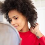 Confident woman with afro hairstyle glancing in the mirror, adjusting her hair indoors, illustrating Natural Hair Care Routine: Products and Techniques for Healthy, Strong Hair