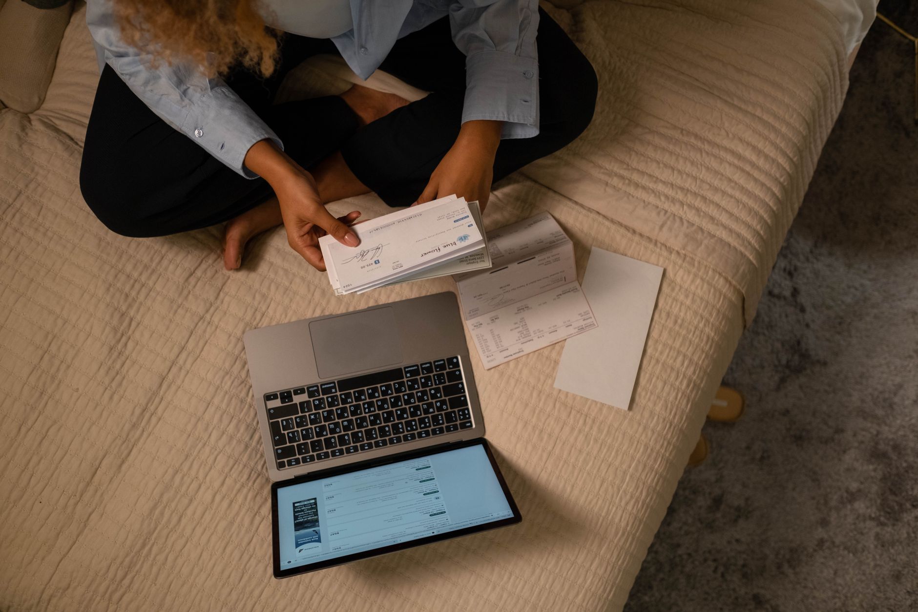 Overhead view of woman organizing finances on bed with laptop and checks, illustrating The bigger leak is the money a no spend month lets you ignore
