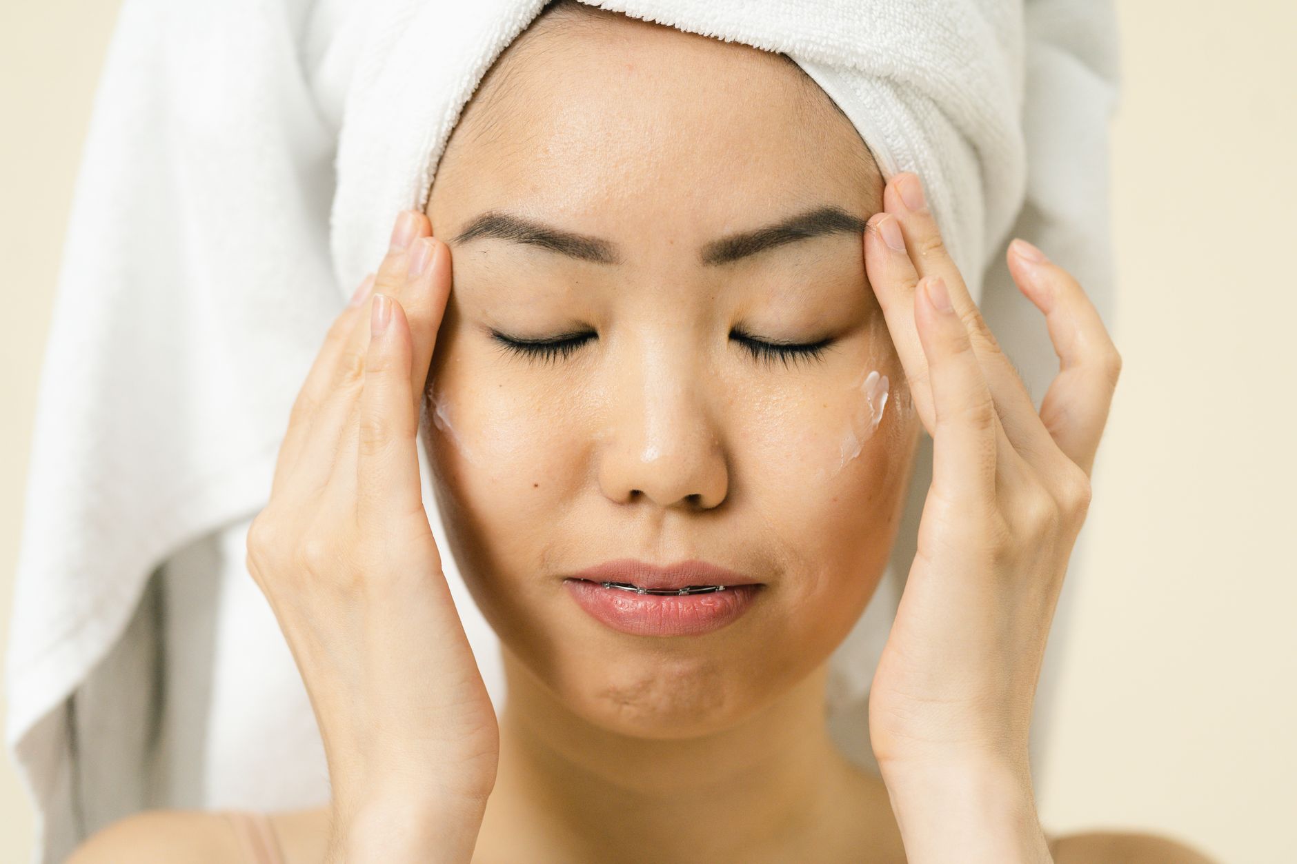 Asian woman with a towel on head applying moisturizer for skincare routine, illustrating Peptides: Building Blocks for Firmer Skin