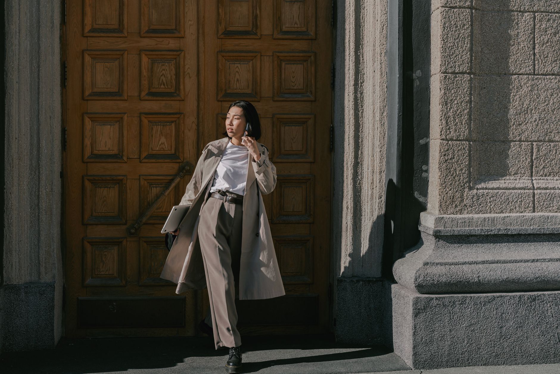 Trench-coated woman standing confidently in front of a wooden door during the day, using a smartphone, illustrating Building Business Casual Outfits That Work Across Seasons