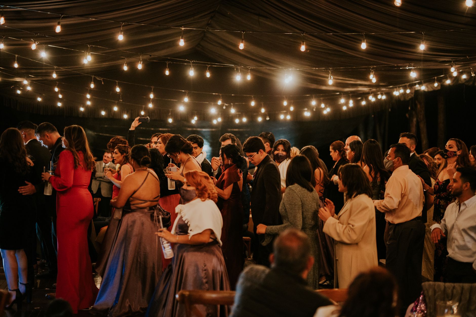 A joyful indoor wedding celebration with elegantly dressed guests under twinkling string lights, illustrating Nobody is tracking your wedding guest dress across weddings