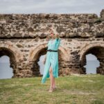 Stylish woman in blue dress posing by Rumelifeneri Castle ruins, illustrating Wedding Guest Dress: Rent or Buy? The Cost Per Wear Math Says Buy (Most of the Time)