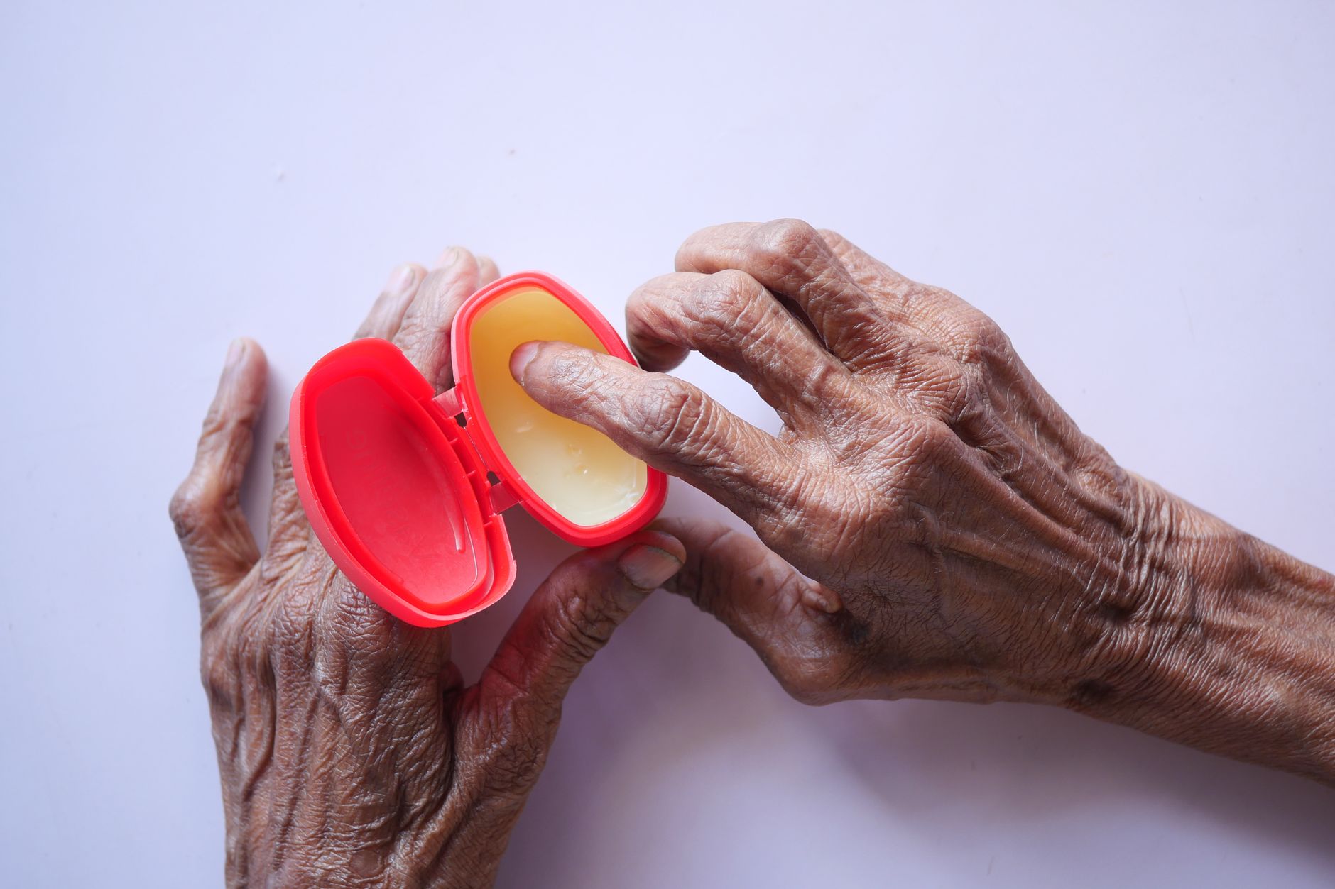 Close-up of senior hands using hand cream for skincare in a red container on a white background, illustrating Then let a simple cream do its job